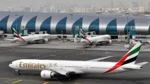 Three Emirates airplanes are parked at a busy airport terminal with a modern, arched design in the background, set against a city skyline.
