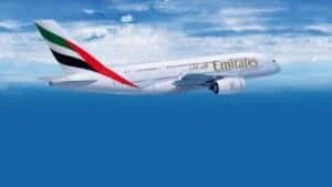 An Emirates Airbus A380 flying in a clear blue sky with white clouds, displaying the airline's logo and flag on the tail.