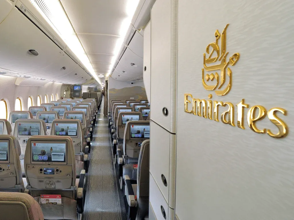 Interior view of an Emirates airplane economy class dubai flights cabin with rows of seats featuring individual entertainment screens, and the Emirates logo on a wall with gold lettering.