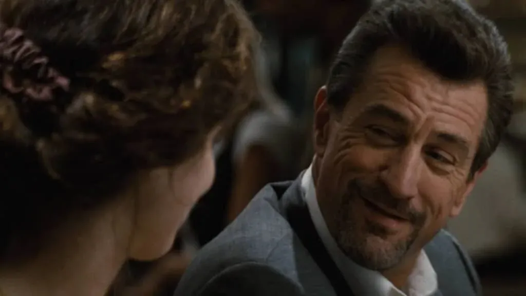 A smiling man in a suit looks at a woman with brown curly hair tied back, in a dimly lit setting.