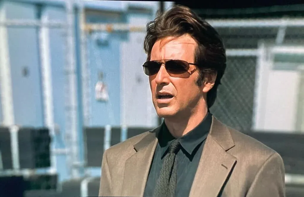A man with dark hair wearing sunglasses and a beige suit stands outdoors, looking away from the camera.