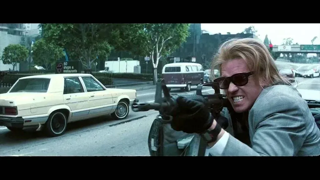 A person with long blond hair and sunglasses holding a rifle aggressively, standing on a city street next to a parked beige car, with other vehicles and trees in the background.