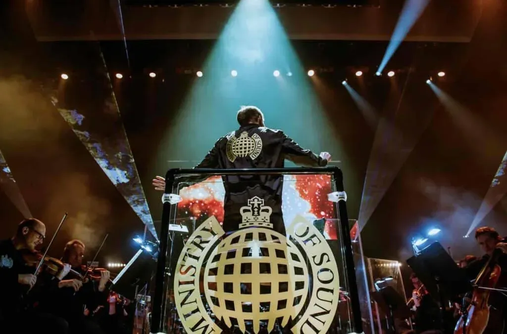A conductor stands on a podium with the Ministry of Sound logo, leading an orchestra under dramatic stage lighting.