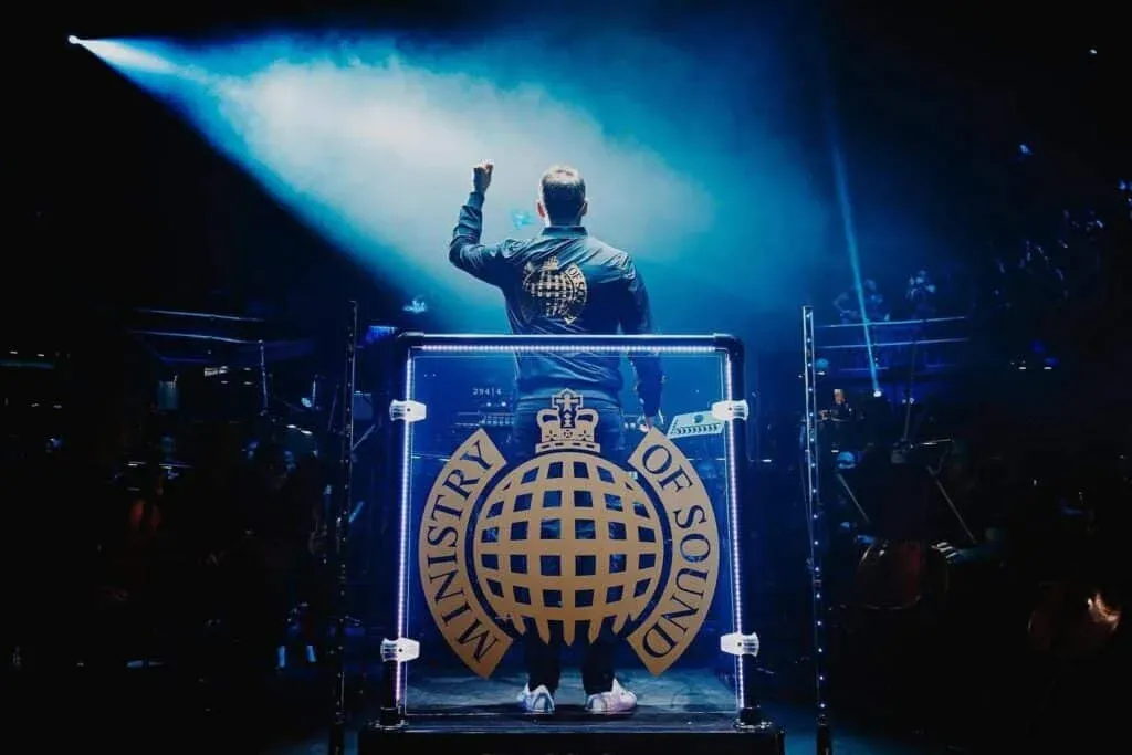 A person standing behind a DJ booth with a large "Ministry of Sound" logo, facing a crowd with raised arm, under dramatic stage lighting.