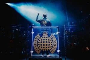A person standing behind a DJ booth with a large "Ministry of Sound" logo, facing a crowd with raised arm, under dramatic stage lighting.