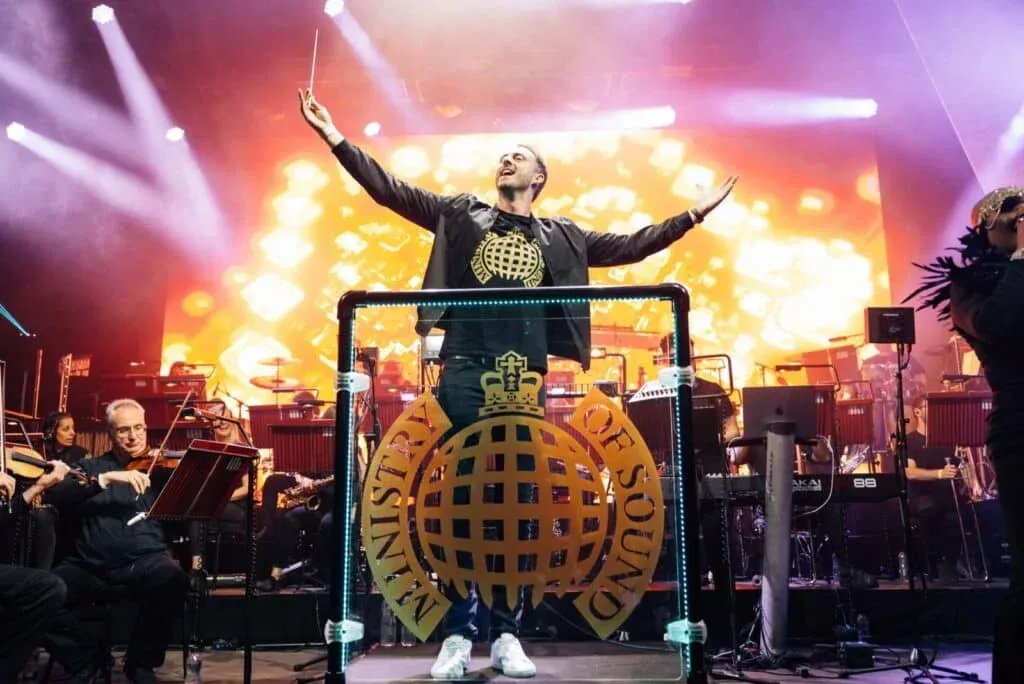 A person on stage conducting an orchestra with the Ministry of Sound logo in front, bathed in colorful stage lighting.