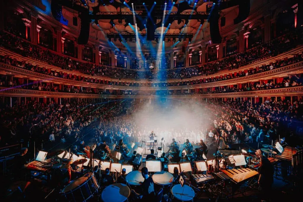 A large orchestra performs on stage in a grand concert hall filled with a large audience. The venue is opulently decorated with multiple tiers of seating, illuminated by dramatic lighting and beams. The conductor is visible in the center, and musicians can be seen with a variety of instruments.