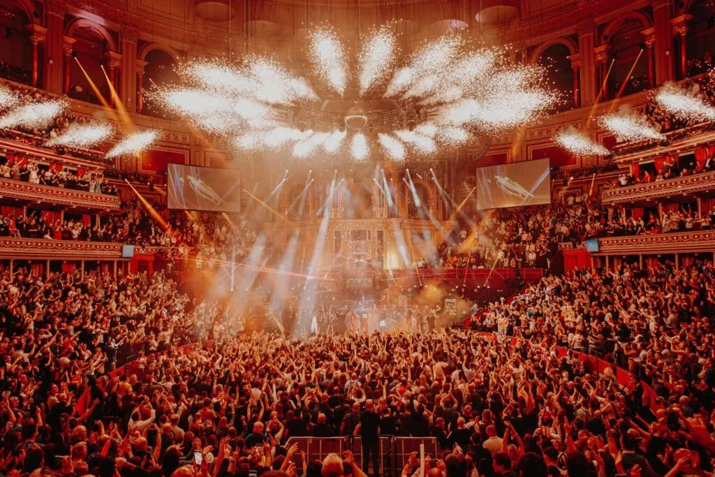 A large, enthusiastic crowd in an ornate concert hall watches a dramatic light show and pyrotechnic display on stage.