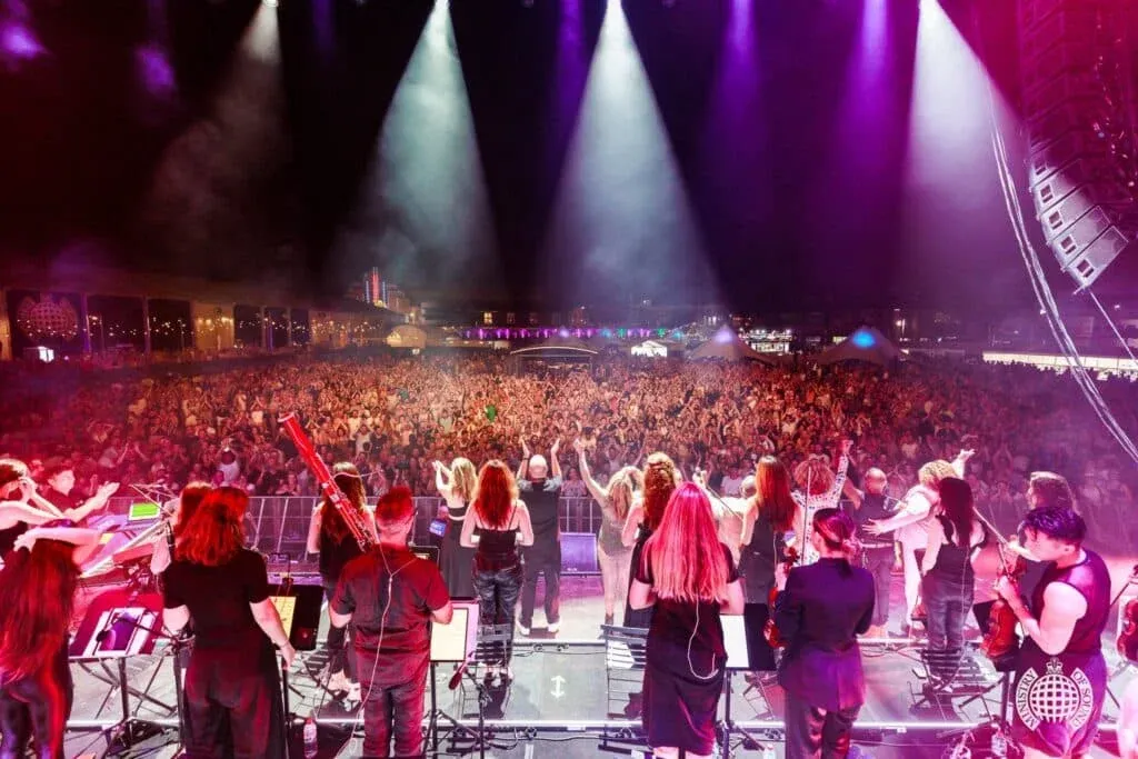 A large orchestra performing on stage with bright spotlights shining down, facing a cheering and packed outdoor audience at night.