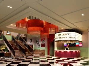 A theater box office with a classic marquee displaying show information for 'Shrek' on Wednesday, March 8, is located in an elegant lobby with a checkered black-and-white marble floor. The space features large red chandeliers with fringe, a wooden staircase, and posters on the walls. A person stands at the box office counter.