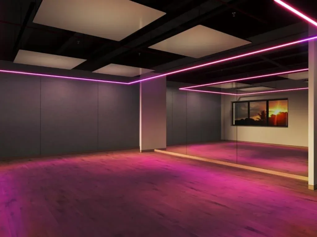 An empty dance studio with wooden flooring and mirrored walls, illuminated by pink neon lights along the ceiling edges.