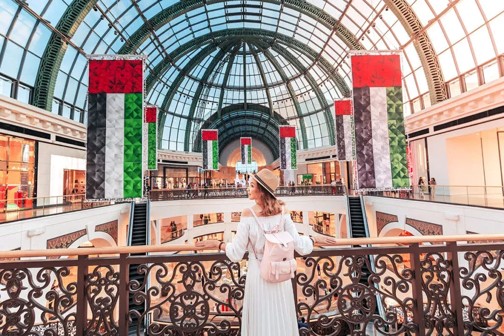 A person wearing a straw hat and a white dress with a pink backpack stands on an ornate balcony inside a large, arched glass dome shopping mall adorned with multiple United Arab Emirates flags.