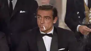 A man in a tuxedo with a bow tie is seated at a table, holding a cigarette in his mouth, surrounded by others in formal attire.