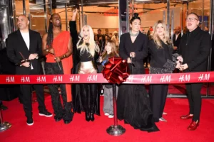 A group of six individuals stands on a red carpet at an H&M store opening, joyfully preparing to cut a large ribbon adorned with a red bow. They are dressed in fashionable attire, with one person holding scissors and others smiling and posing with excitement.