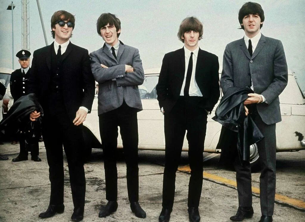 Four men in suits stand on a concrete surface in front of a white car, with one wearing sunglasses and holding a coat, while two others also hold coats.