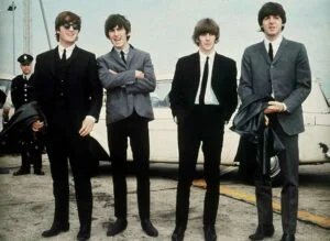 Four men in suits stand on a concrete surface in front of a white car, with one wearing sunglasses and holding a coat, while two others also hold coats.