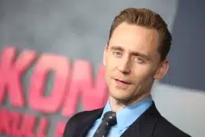 A person in formal attire with a suit and tie, standing in front of a blurred background featuring the letters 'KON'.