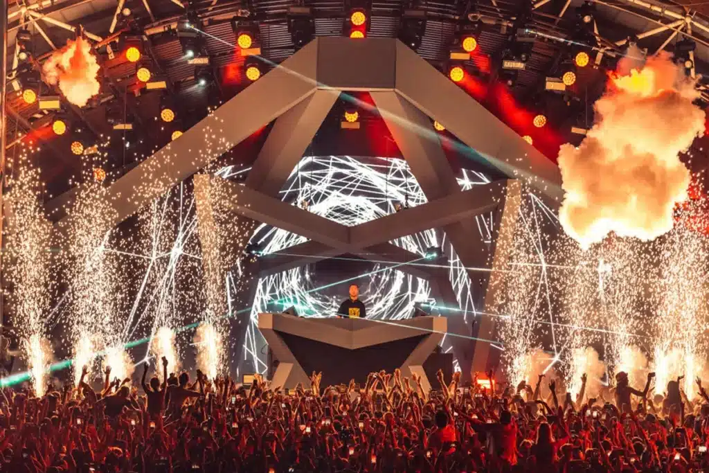 A DJ performs at a lively music festival with a large crowd cheering in front of a futuristic stage lit with vibrant red and orange lighting, surrounded by dynamic pyrotechnics and visual effects on a large screen backdrop.