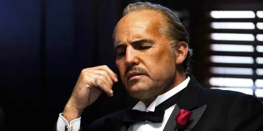 A man in formal attire with a serious expression and closed eyes, wearing a tuxedo with a red rose boutonniere, seated indoors near a window with blinds.