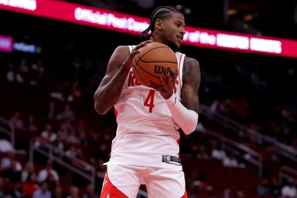 A basketball player in a white and red uniform holding a basketball on the court, with the stands visible in the background.