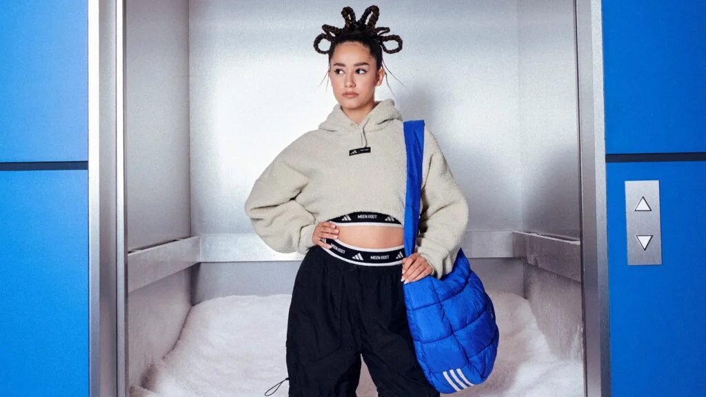 A person with unique coiled hairstyle stands confidently in an elevator, wearing a beige cropped hoodie and black pants, with a blue quilted bag over one shoulder.