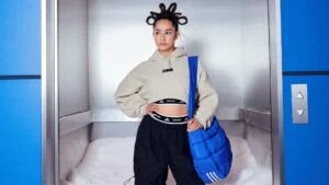 A person with unique coiled hairstyle stands confidently in an elevator, wearing a beige cropped hoodie and black pants, with a blue quilted bag over one shoulder.