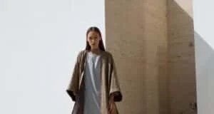 Person wearing a flowy, light-colored outfit with a long, open coat, standing in front of a minimalist background with angular walls and soft lighting.