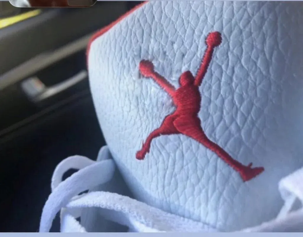 Close-up of a sneaker with a red Jumpman logo embroidered on textured white leather, featuring white laces.
