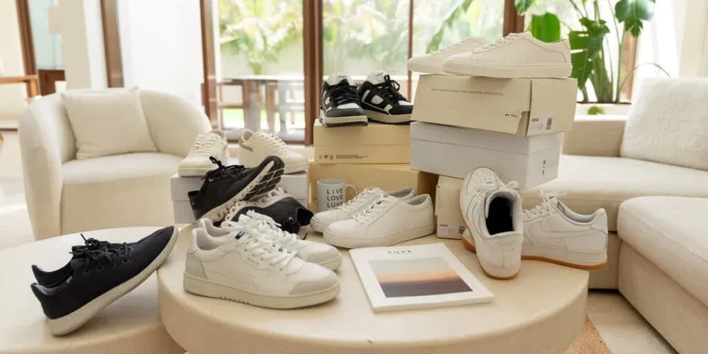A variety of sneakers in black and white are displayed on a circular table alongside several shoe boxes in a modern living room with a beige armchair and large windows.