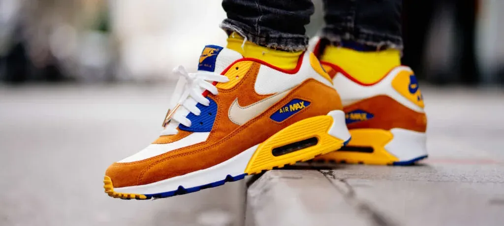 A stylish pair of Nike Air Max sneakers in orange, white, and blue, with a visible air cushion in the heel, worn by a person standing on a concrete edge.