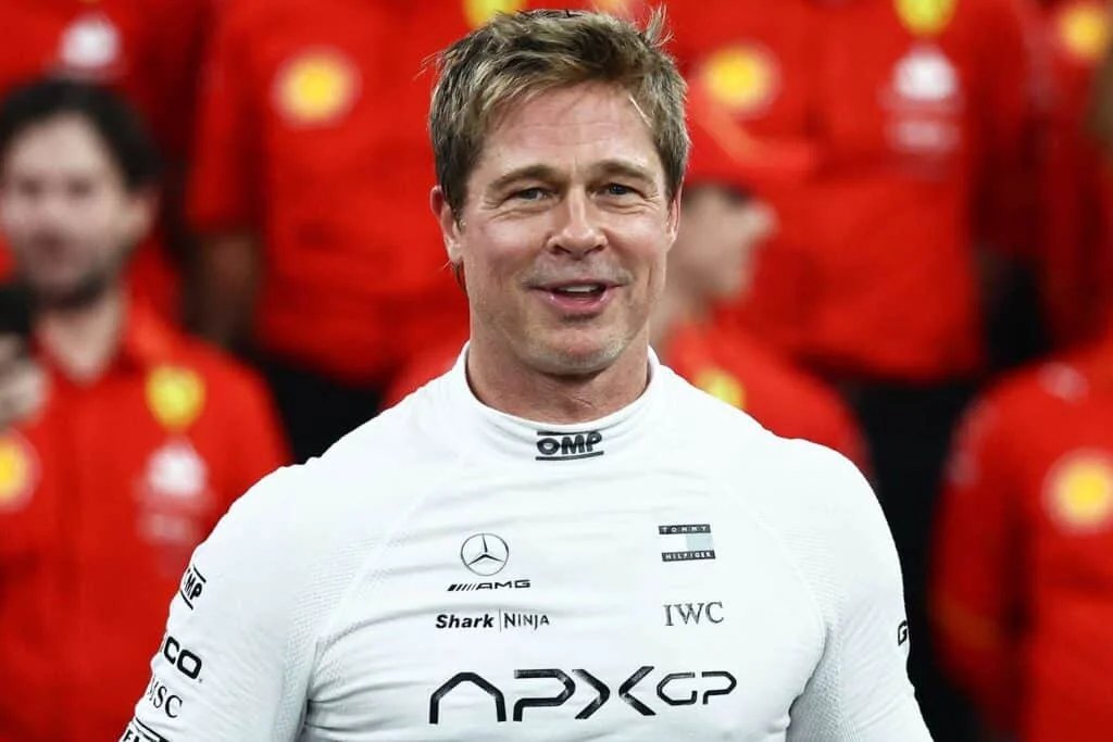 A man wearing a white racing suit with various logos stands smiling in front of a group of people wearing red uniforms.