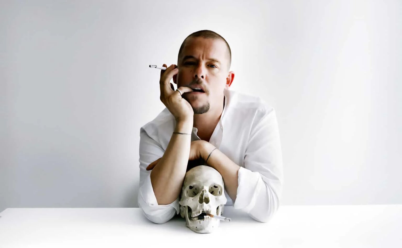 A person in a white shirt leans on a table holding a cigarette, with a skull positioned in front of them also holding a cigarette.