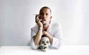 A person in a white shirt leans on a table holding a cigarette, with a skull positioned in front of them also holding a cigarette.