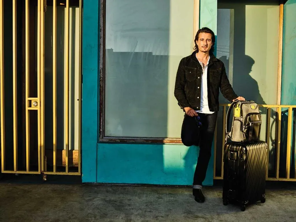 A person in casual attire, wearing a black jacket and jeans, leans against a suitcase in front of a building with blue walls and yellow railing, lit by sunlight.