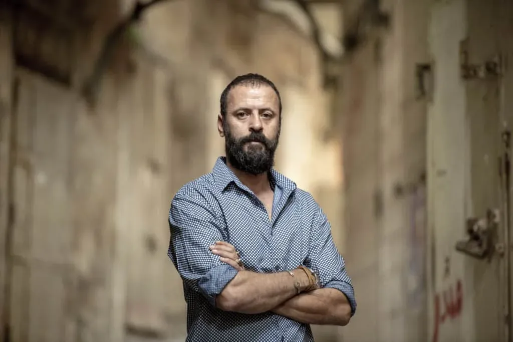 A man with a beard and short hair stands with his arms crossed in a narrow, blurred corridor, wearing a patterned blue shirt.