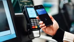 A person holding a smartphone with a digital wallet app displaying a credit card, preparing to use contactless payment at a card reader.