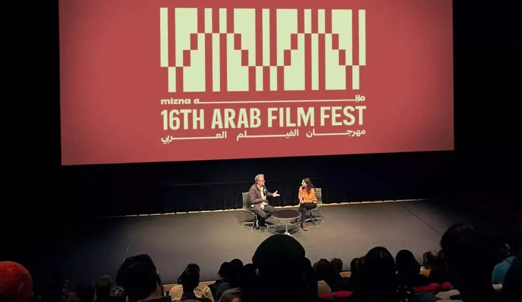 Two people sit on a stage in front of an audience at the 16th Arab Film Fest, with a large event banner displayed above them.
