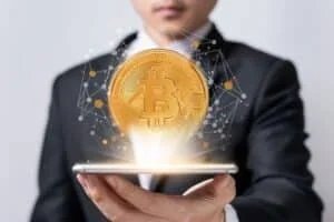 A person in a suit is holding a smartphone, with a large digital representation of a Bitcoin coin hovering above the screen, surrounded by abstract network connections and glowing light effects.