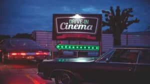 Two vintage cars in front of a lit-up 'Drive-In Cinema' sign at dusk, with arrows indicating the direction for the cinema entrance.