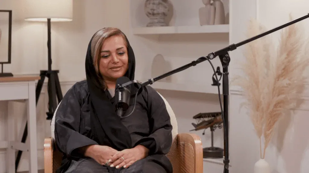 A person wearing a black hijab and robe is seated in front of a microphone in a cozy room with shelves and a lamp in the background.