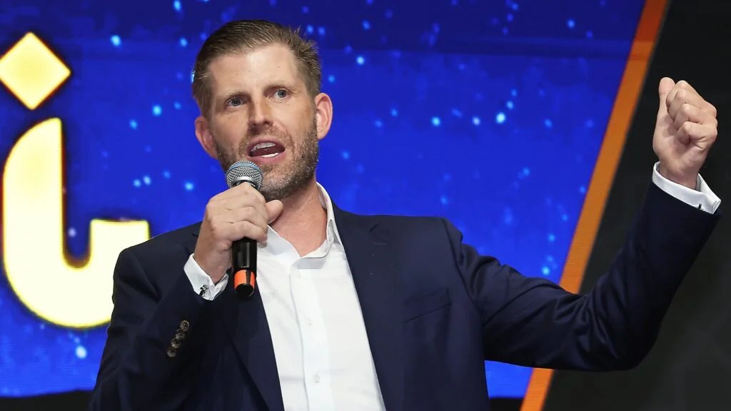 A man in a suit holds a microphone and gestures with his fist raised, speaking in front of a blue background with star-like patterns.