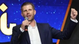 A man in a suit holds a microphone and gestures with his fist raised, speaking in front of a blue background with star-like patterns.