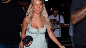 A woman with long blonde hair, wearing a light green dress, smiling while walking at night, surrounded by others.