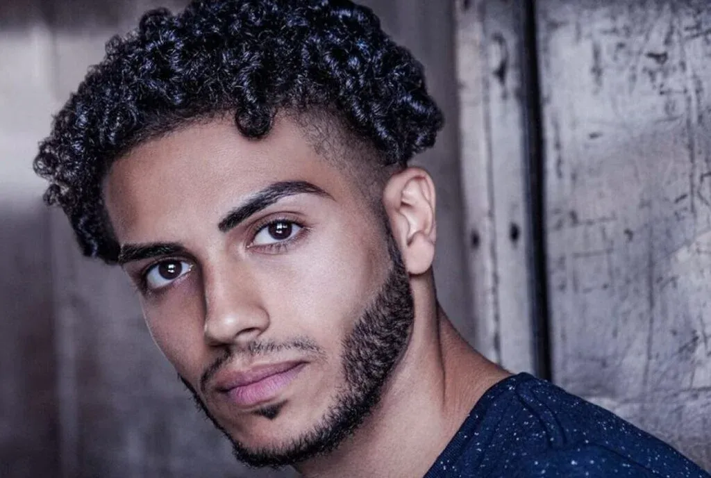 A person with curly hair and a neatly trimmed beard is looking intently at the camera, set against a rustic wooden backdrop.