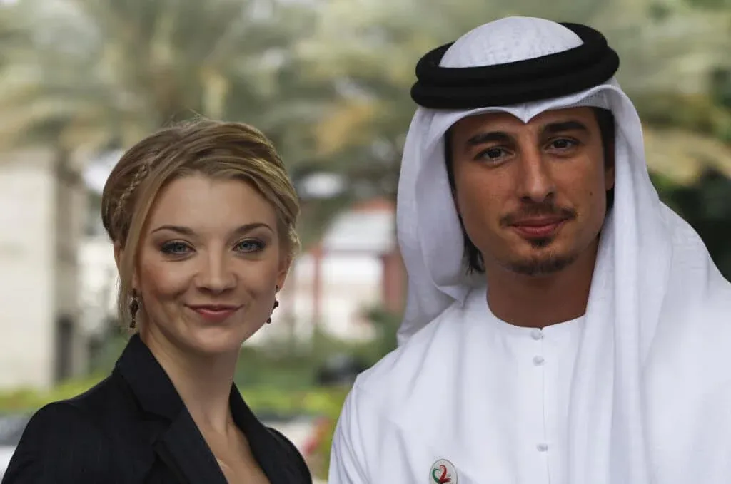 A woman with blonde hair wearing a dark jacket stands next to a man in traditional Middle Eastern attire, featuring a white kandura and black agal. They are outdoors with a blurred background of trees.
