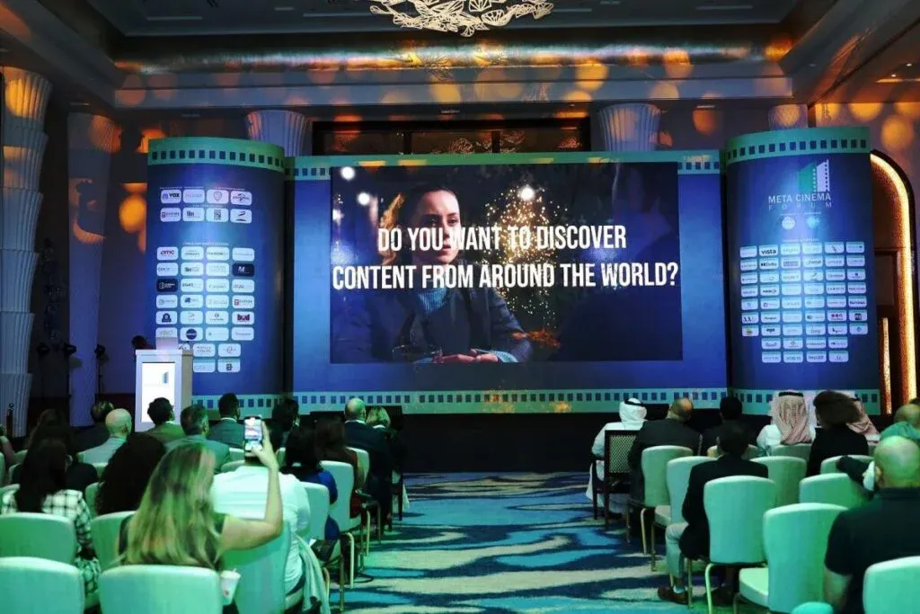 Audience seated in a conference room facing a large screen displaying the text 'Do you want to discover content from around the world?' with conference branding in the background.