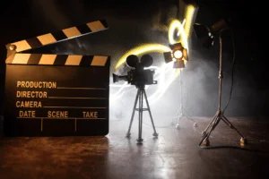 A film production setup with a clapperboard in the foreground, an old-fashioned movie camera on a tripod in the center, and two studio lights on tripods, all set against a smoky background with bright lights creating streaks.