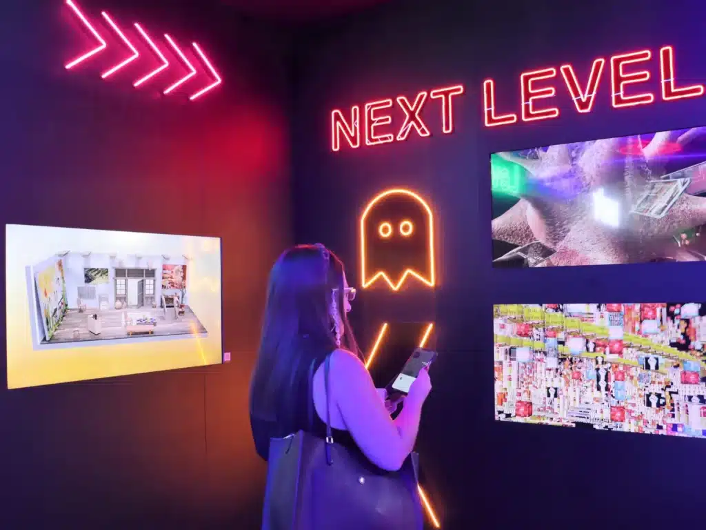 A woman stands in a dimly lit room with neon lights and digital art displays. The wall features the words 'NEXT LEVEL' in bright pink neon, alongside an outline of a ghost-like figure. The room is illuminated with colorful lights highlighting various digital artworks.