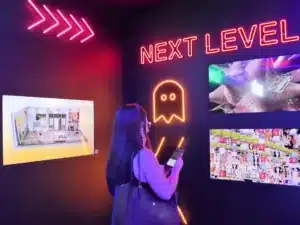 A woman stands in a dimly lit room with neon lights and digital art displays. The wall features the words 'NEXT LEVEL' in bright pink neon, alongside an outline of a ghost-like figure. The room is illuminated with colorful lights highlighting various digital artworks.