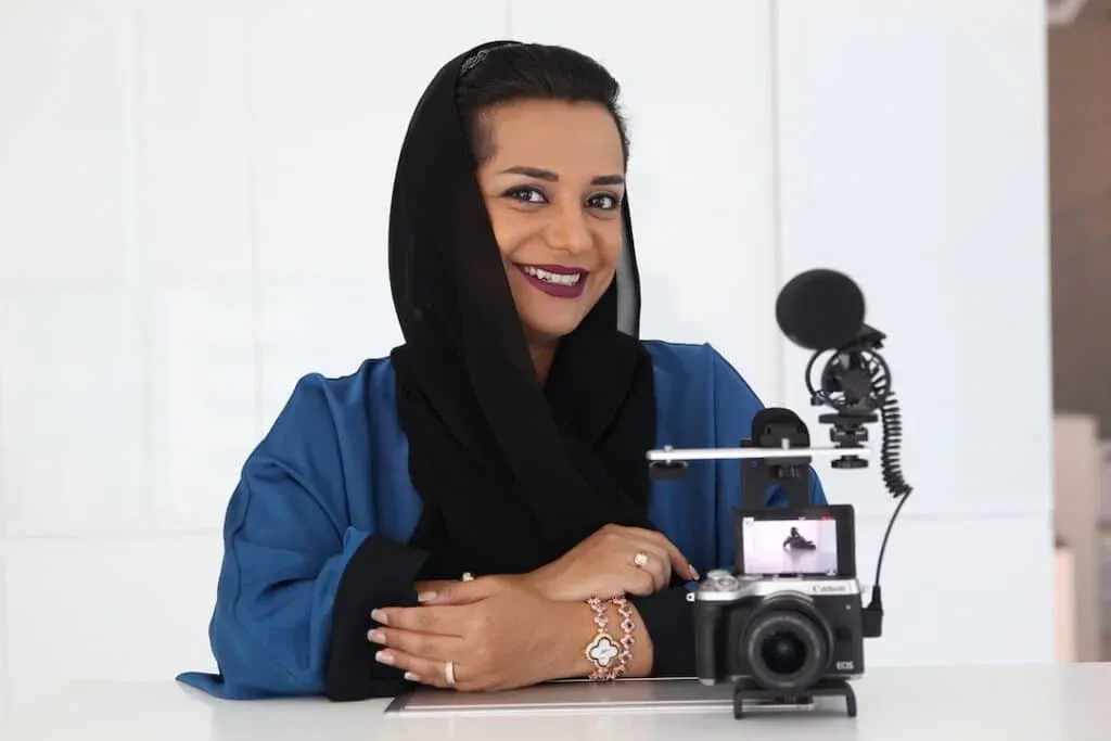 A woman wearing a blue top and black hijab smiles while sitting behind a professional camera with a microphone attached.